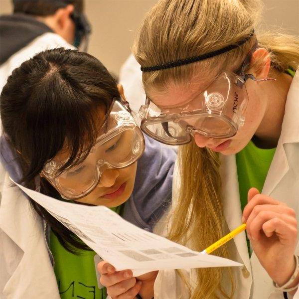 Two people wearing goggles peer at a piece of paper that one person is holding. That person is also holding a pencil.