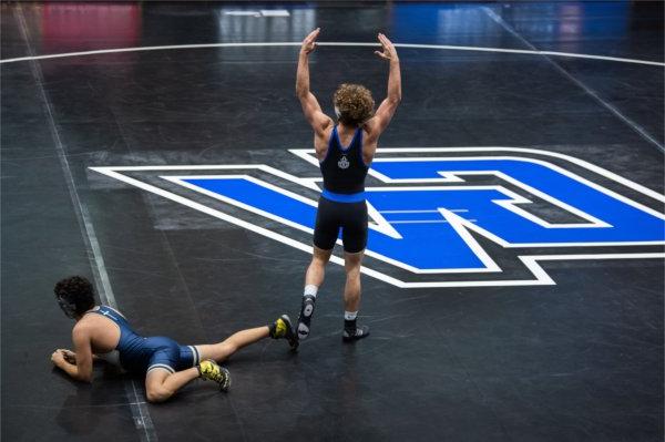 一个人 stands on the mat with his hands in the air in victory after a wrestling match while his opponent is still on the ground. 