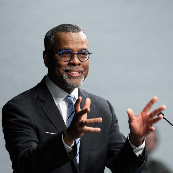 Eddie Glaude Jr. at podium, raising hands to audience. He is wearing a dark suit and blue framed glasses